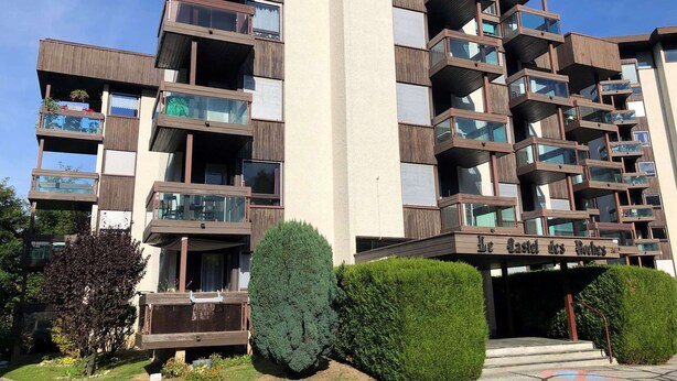 Un immeuble résidentiel à plusieurs étages, doté de balcons, d’une enseigne indiquant « Maison des Voleurs » et d’un aménagement paysager bien entretenu.