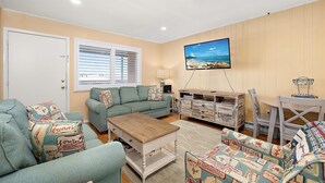 A living room with a sofa, a coffee table, a TV mounted on the wall, and a dining area with a table and chairs. - Oceanside Three Bedroom Condo Steps From the Beach (Ocean City)