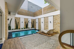 A modern outdoor pool area with a stone wall, a lounge chair, and a circular woven table. - Gloen Villa by Betterplace (Canggu)