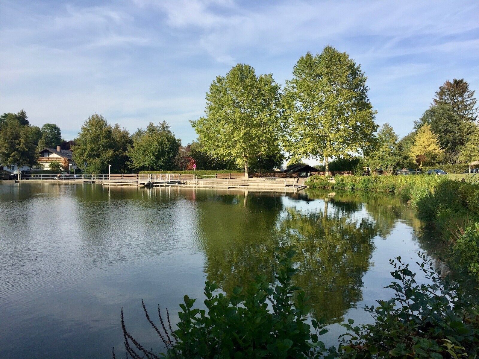 A calm lake with a wooden bridge, surrounded by trees and a building in the background.