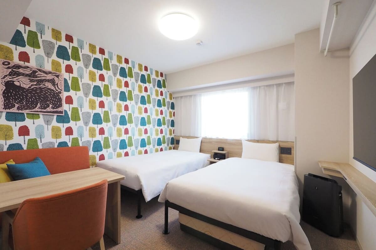 Habitación de hotel con dos camas, un escritorio, una silla, una pared colorida, una ventana con cortinas y un televisor.