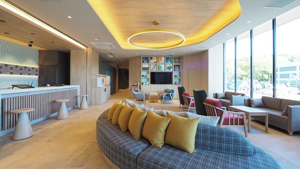 A modern hotel lobby with a large seating area, a reception desk, and a large window offering a view of the outside.