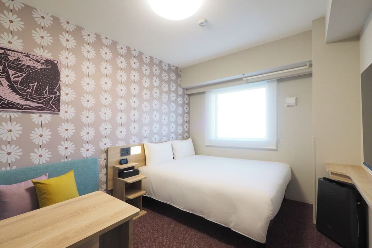 Habitación de hotel con cama, escritorio, silla, pequeña mesa, mesita de noche, lámpara, ventana y pared con estampado floral.