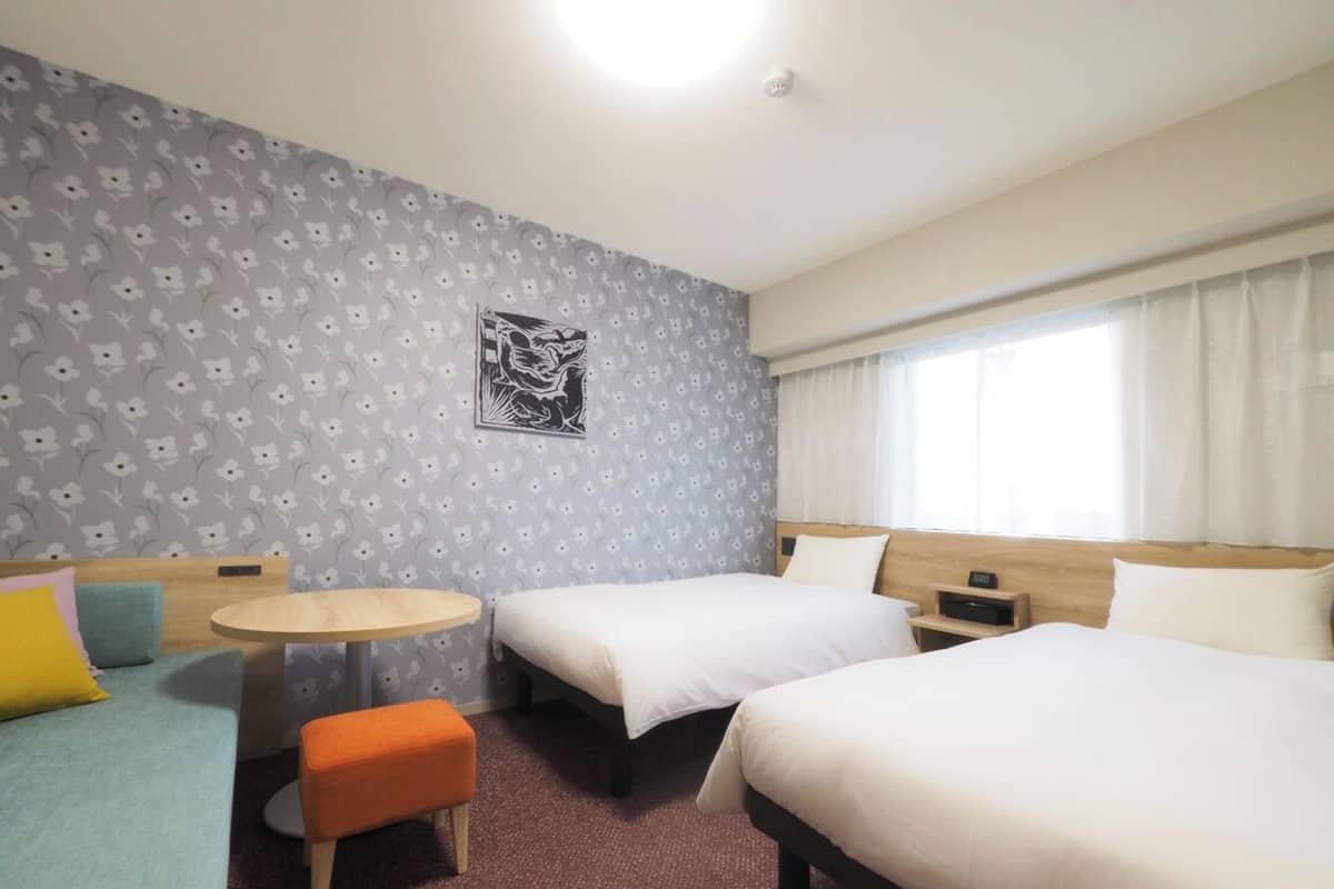 Habitación de hotel con dos camas, una mesita redonda pequeña, un sofá y un papel tapiz estampado.