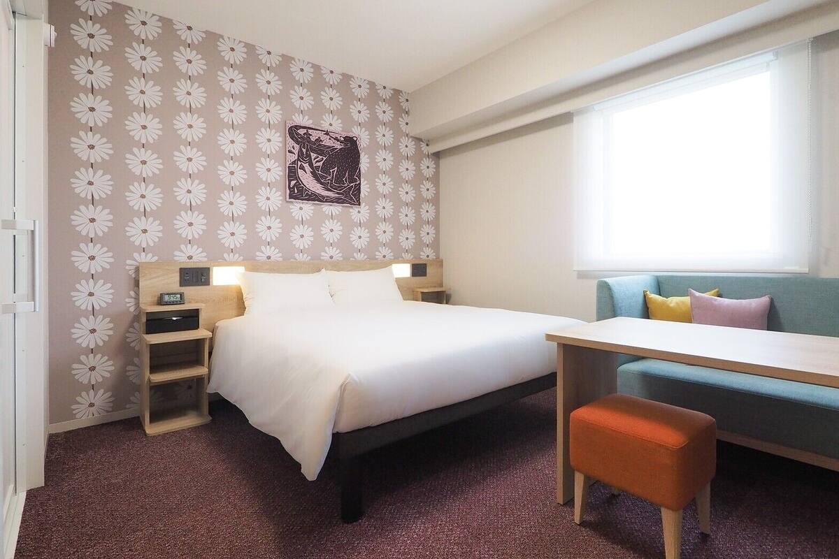 Una habitación de hotel con cama, escritorio, silla, sofá, un reloj de pared y una ventana.