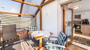 Una terraza de madera con una mesa y sillas, una zona de cocina con armarios y electrodomésticos, y vistas a través de un ventanal.