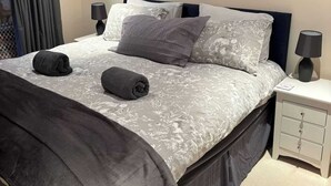 A neatly made bed with a grey duvet and pillows, a bedside table with a lamp, and a framed picture on the wall.