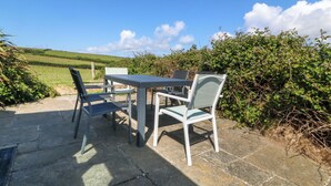 An outdoor patio with a table and chairs, stone flooring, and a view of a green field and hills in the background.