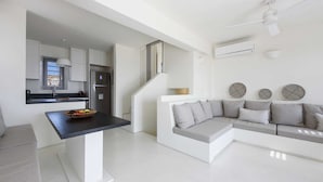 Una sala de estar moderna con un sofá gris, una pequeña mesa de comedor y una zona de cocina con refrigerador y ventana.