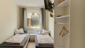 A small room with two beds, a window with curtains, a radiator, and a wardrobe with hangers. - Apartment 'Harzblick Am Waldesrand' with Mountain View, Private Terrace (and Sauna) (Hahausen)