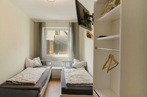 A small room with two beds, a window with curtains, a radiator, and a wardrobe with hangers. - Apartment 'Harzblick am Waldesrand' with Mountain View, Private Terrace & Sauna (Hahausen)