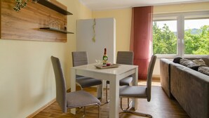 A dining area with a white table and four grey chairs, a grey sofa, and a window with a view of greenery. - Apartment 'Harzblick Am Waldesrand' with Mountain View, Private Terrace (and Sauna) (Hahausen)
