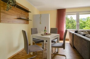 A dining area with a white table and four grey chairs, a grey sofa, and a window with a view of greenery. - Apartment 'Harzblick am Waldesrand' with Mountain View, Private Terrace & Sauna (Hahausen)