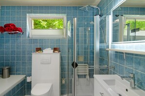 Casa de banho com azulejos azuis, cabine de chuveiro em vidro, lavatório branco e janela com vista para vegetação.