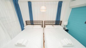 Un lit double avec du linge de lit blanc et une tête de lit en bois, une table de chevet avec une lampe, et une fenêtre avec des rideaux bleus.