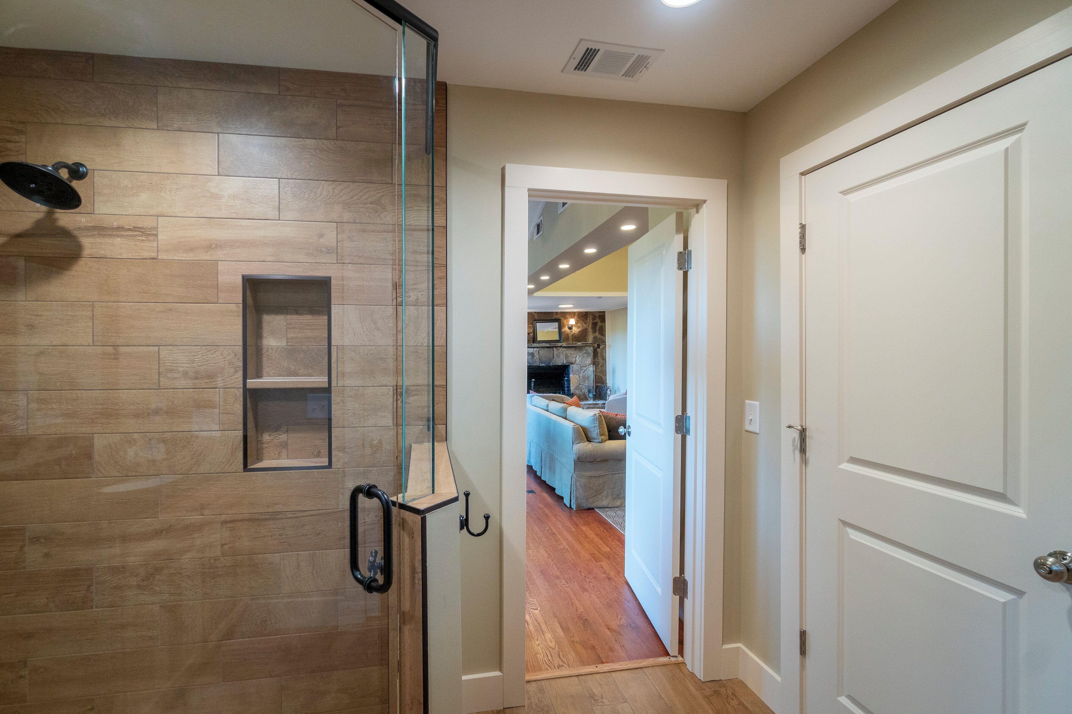 Un couloir doté d’un plancher en bois, d’une cabine de douche en verre et d’une porte blanche donnant sur un espace de vie avec une cheminée.