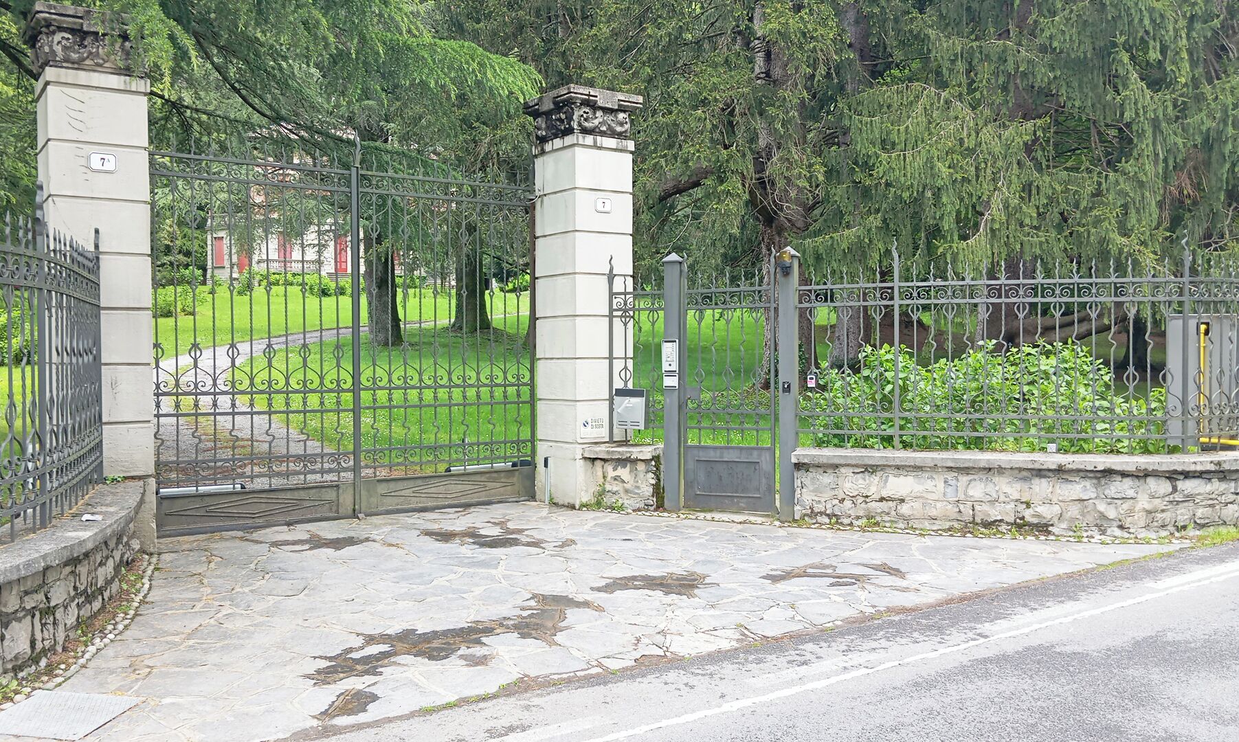 Una entrada cerrada con un pilar de piedra y una verja de metal, rodeada de follaje y árboles.
