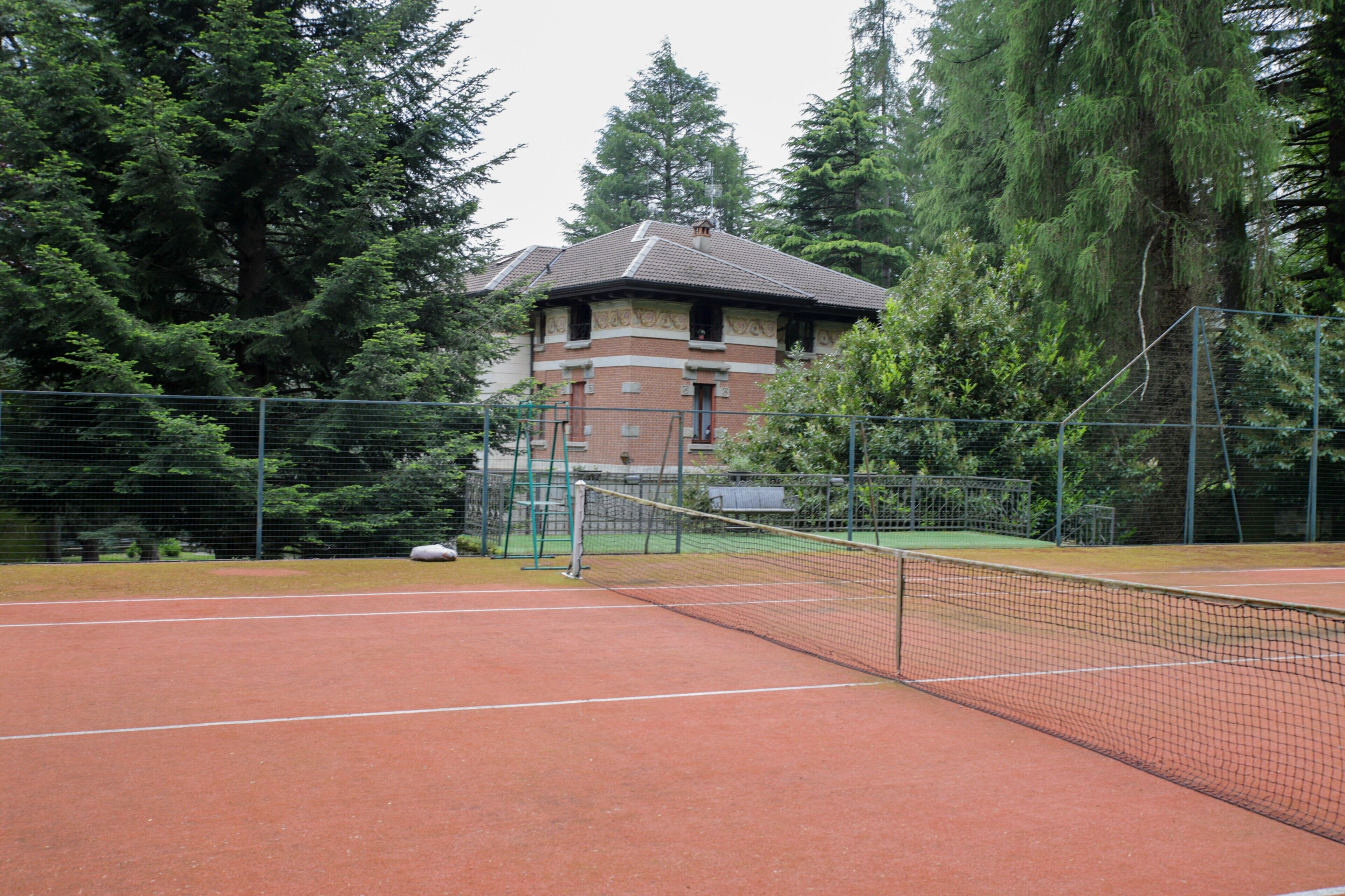 A tennis court with a net, surrounded by a fence, and a building with a brick facade in the background.