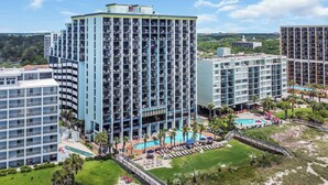 A high-rise hotel with multiple balconies, a swimming pool, and a landscaped area with palm trees.