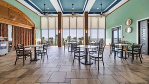 A spacious dining area with multiple tables and chairs, large windows offering a view of palm trees, and a ceiling fan.