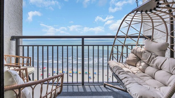 A balcony with a wicker chair and a cushioned bench overlooking a beach with umbrellas and people.