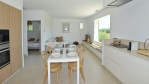 Eine moderne Küche mit weißen Schränken, ein Essbereich mit weißem Tisch und Stühlen sowie ein großes Fenster mit Blick ins Freie.