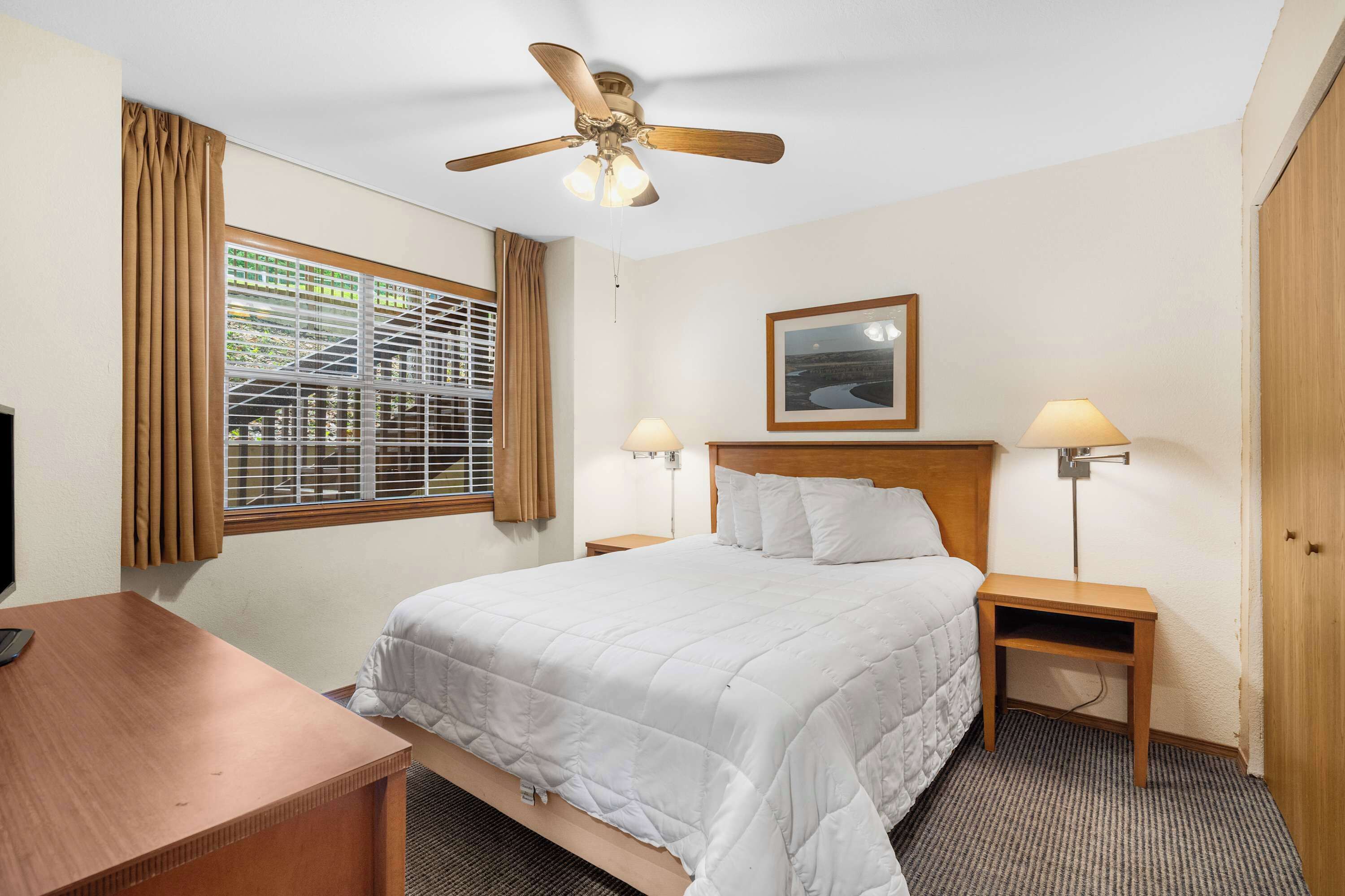 A hotel room with a bed, a desk, a ceiling fan, two bedside lamps, a framed picture on the wall, and a window with curtains.