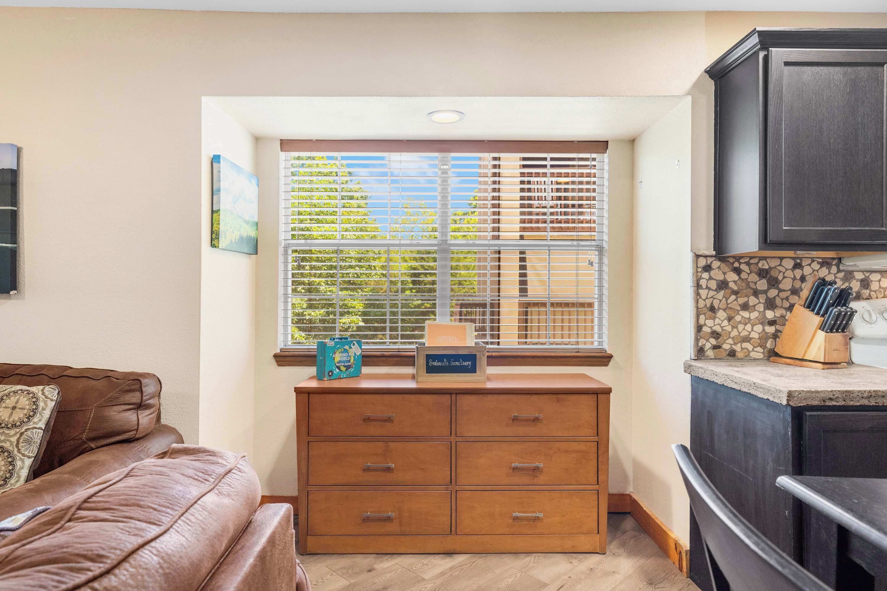 A corner of a room with a brown leather sofa, a wooden cabinet with drawers, a window with blinds, and a kitchen area with a stone backsplash and knife block.