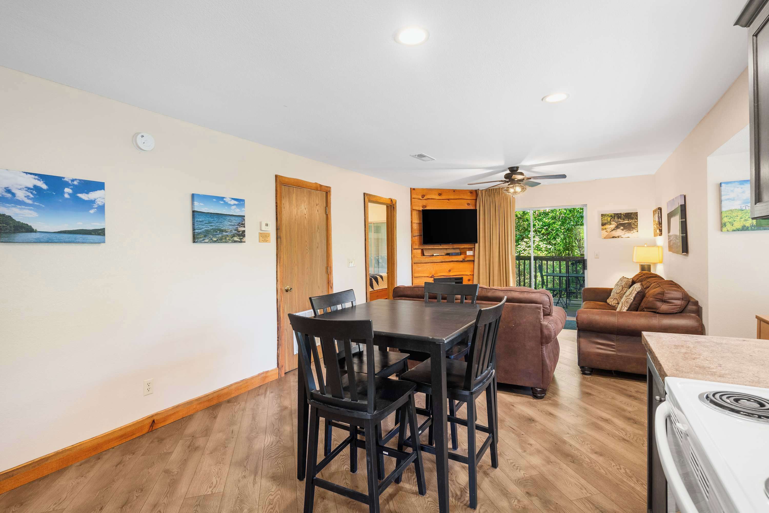 A dining area with a table and chairs, a sofa, a TV mounted on a wooden panel, and a ceiling fan.
