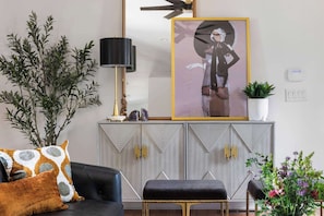 Una sala de estar con sofá, reposapiés, planta en maceta, una obra de arte enmarcada y una lámpara decorativa.