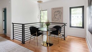 A modern dining area with a round table, two black chairs, and a black railing. A large abstract painting is on the wall, and there is a window with a view of buildings outside.