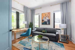 Een moderne woonkamer met een grijze bank, een blauwe fauteuil, een glazen salontafel met een groene kom, en een schilderij van vogels aan de muur.