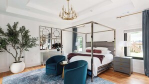 A modern bedroom with a four-poster bed, two armchairs, a large potted plant, and a view of the outdoors.