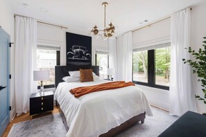 Ein Schlafzimmer mit einem großen Bett, einem Nachttisch, einer Lampe, einem Fenster mit Vorhängen und Blick ins Freie.