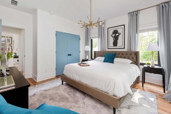 A bedroom with a large bed, a desk, a chair, a window with curtains, and a chandelier.