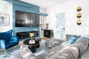 A modern living room with a blue sofa, a grey sectional, a black coffee table, and a flat-screen TV mounted on the wall.