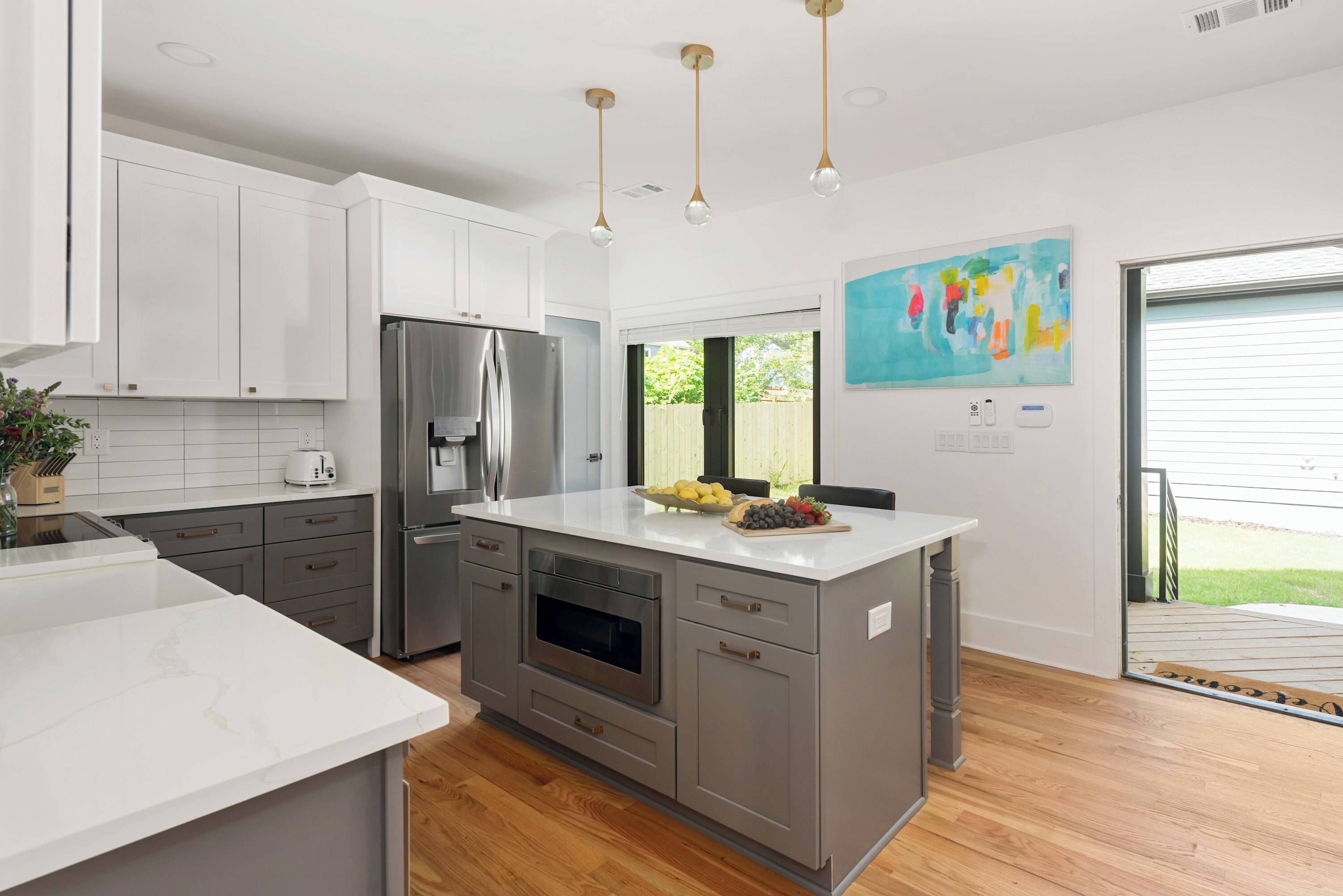 A modern kitchen with white cabinets, a stainless steel refrigerator, an oven, and a large island with a bowl of fruit.