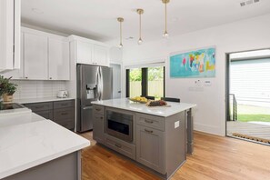 A modern kitchen with white cabinets, a stainless steel refrigerator, an oven, and a large island with a bowl of fruit.