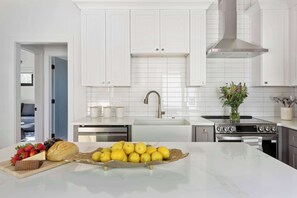 Une cuisine moderne avec des meubles blancs, une hotte en acier inoxydable et un îlot central agrémenté d’un panier de fruits.