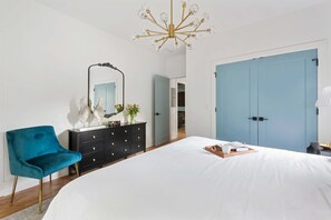 Une chambre à coucher avec un grand lit, une armoire bleue, une commode noire, un fauteuil turquoise et un lustre.