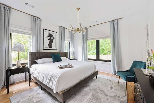 A bedroom with a large bed, a desk, and a chair. There are windows with curtains, a chandelier, and a framed picture on the wall.