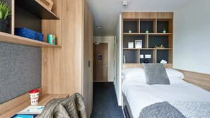 Una habitación de hotel con una cama, un escritorio con un cuaderno, una taza y un estante con una planta y una caja.