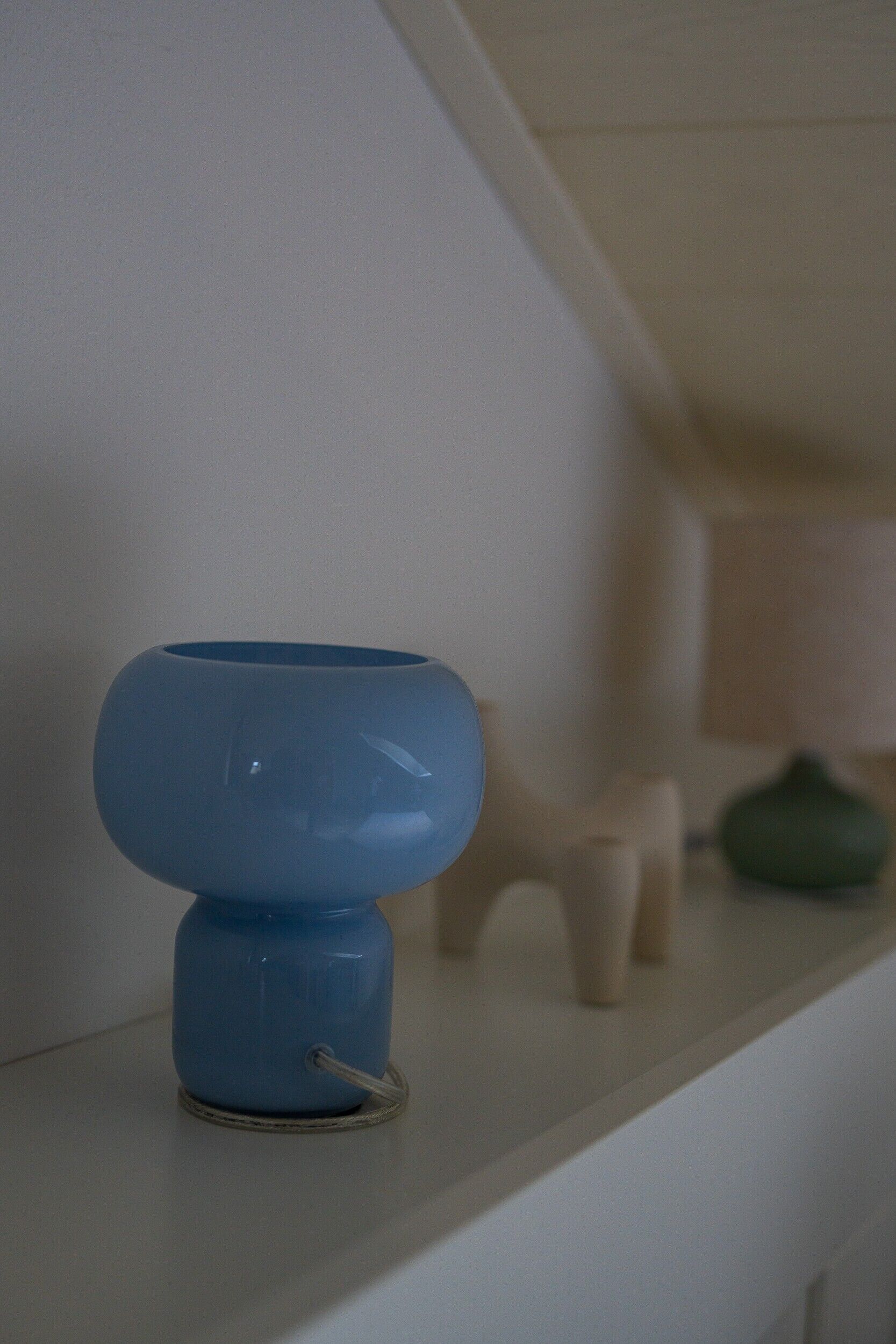 Um objeto azul numa prateleira branca, com uma lâmpada e uma pequena figura no fundo.
