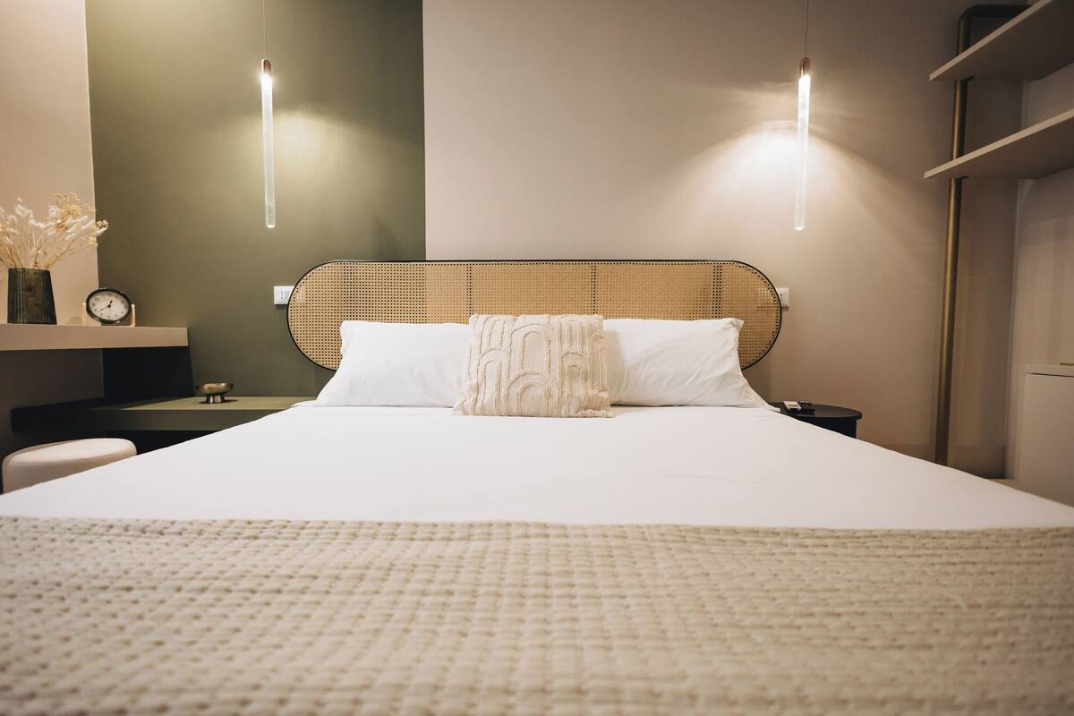 A neatly made bed with a textured blanket, a bedside table with a clock, and a wall-mounted light fixture.