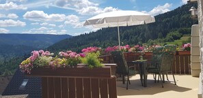 A terrace with a table and chairs, a white umbrella, and a flower box with various plants.