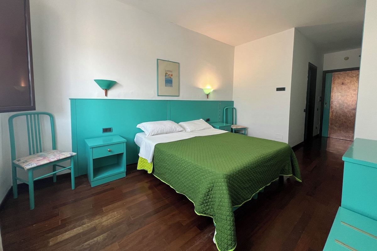 A bedroom with a bed, a nightstand, a chair, a wall-mounted lamp, and a door.