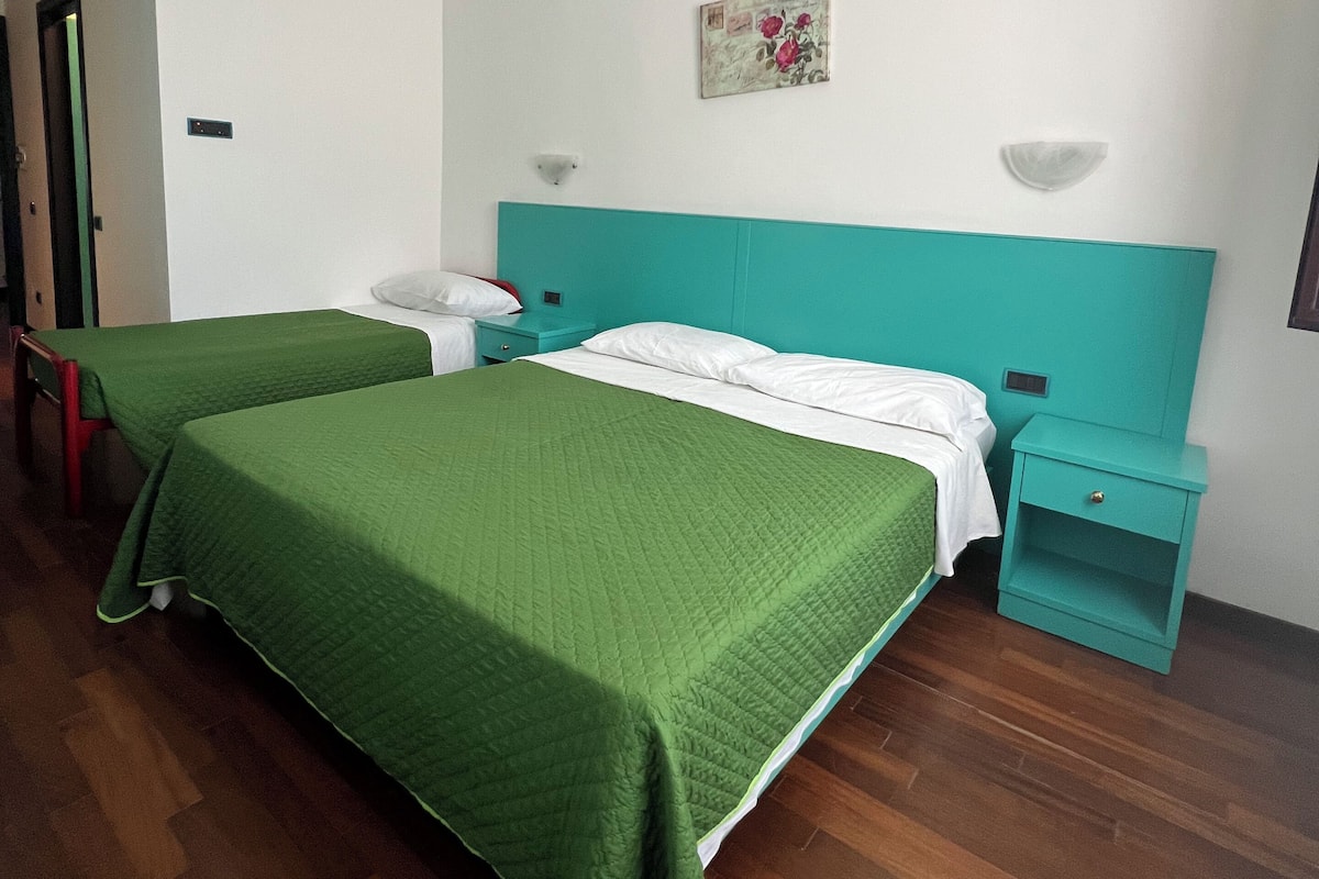 A room with two beds, each with a green bedspread, a teal headboard, a small bedside table, and a wall-mounted light.