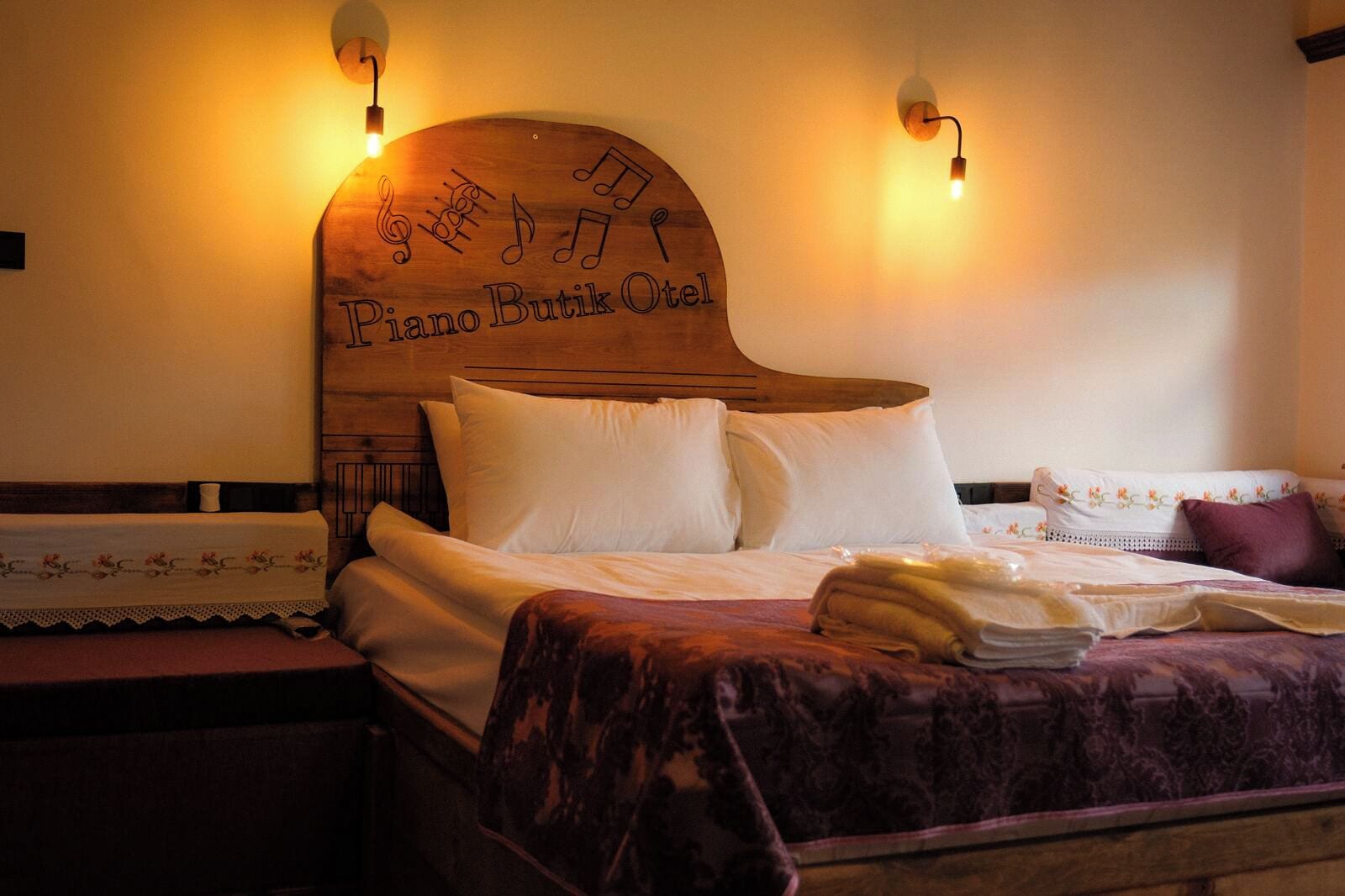 Ein Hotelzimmer mit einem Holzkopfteil, verziert mit Noten und dem Schriftzug "Piano Butik Oslo". Zwei Nachttischlampen, ein Bett mit weißen Leinen und eine violette Tagesdecke sind ebenfalls vorhanden.