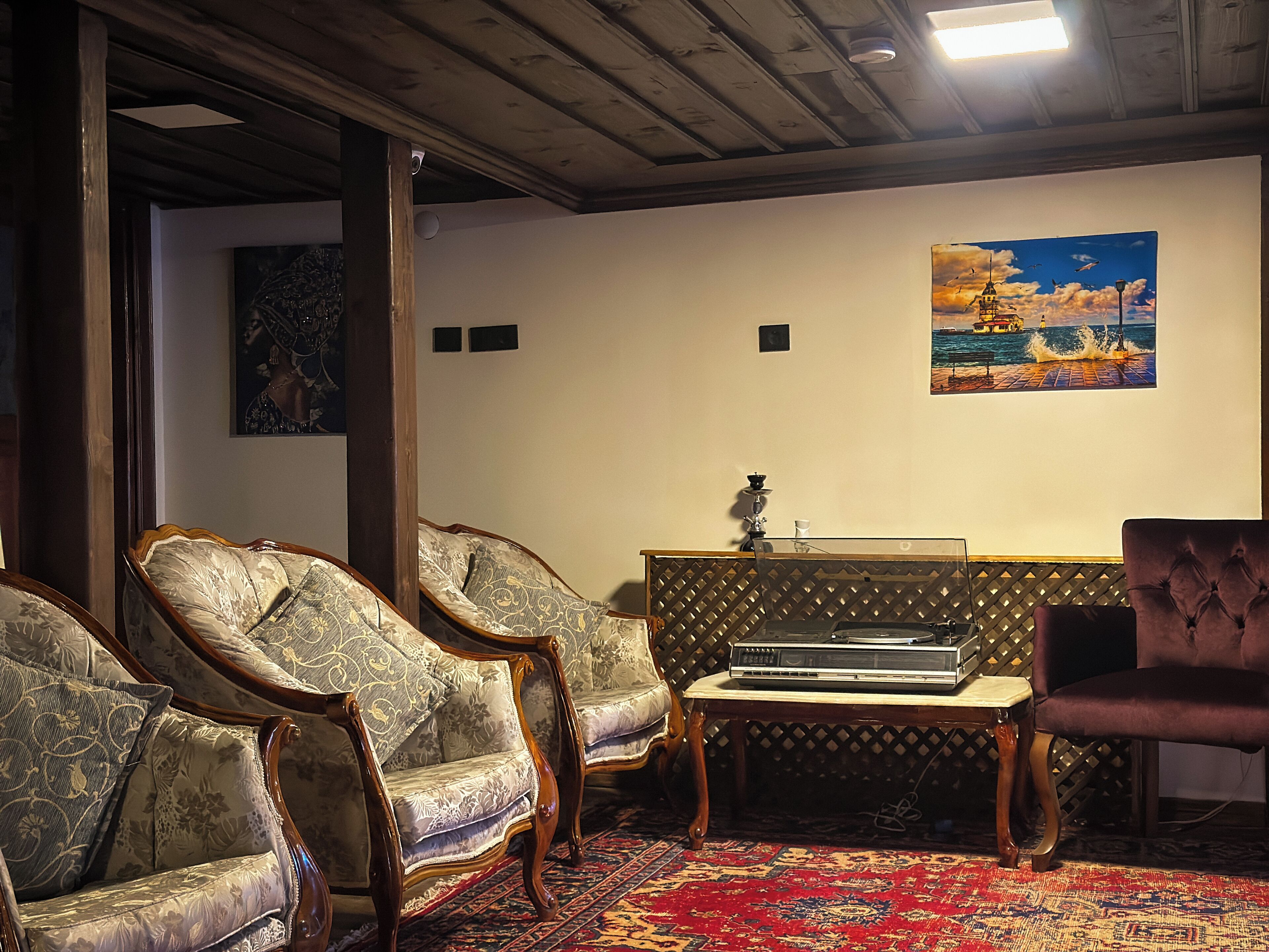 A room with three upholstered chairs, a small table with a stereo system, a patterned rug, and a painting of a coastal scene.