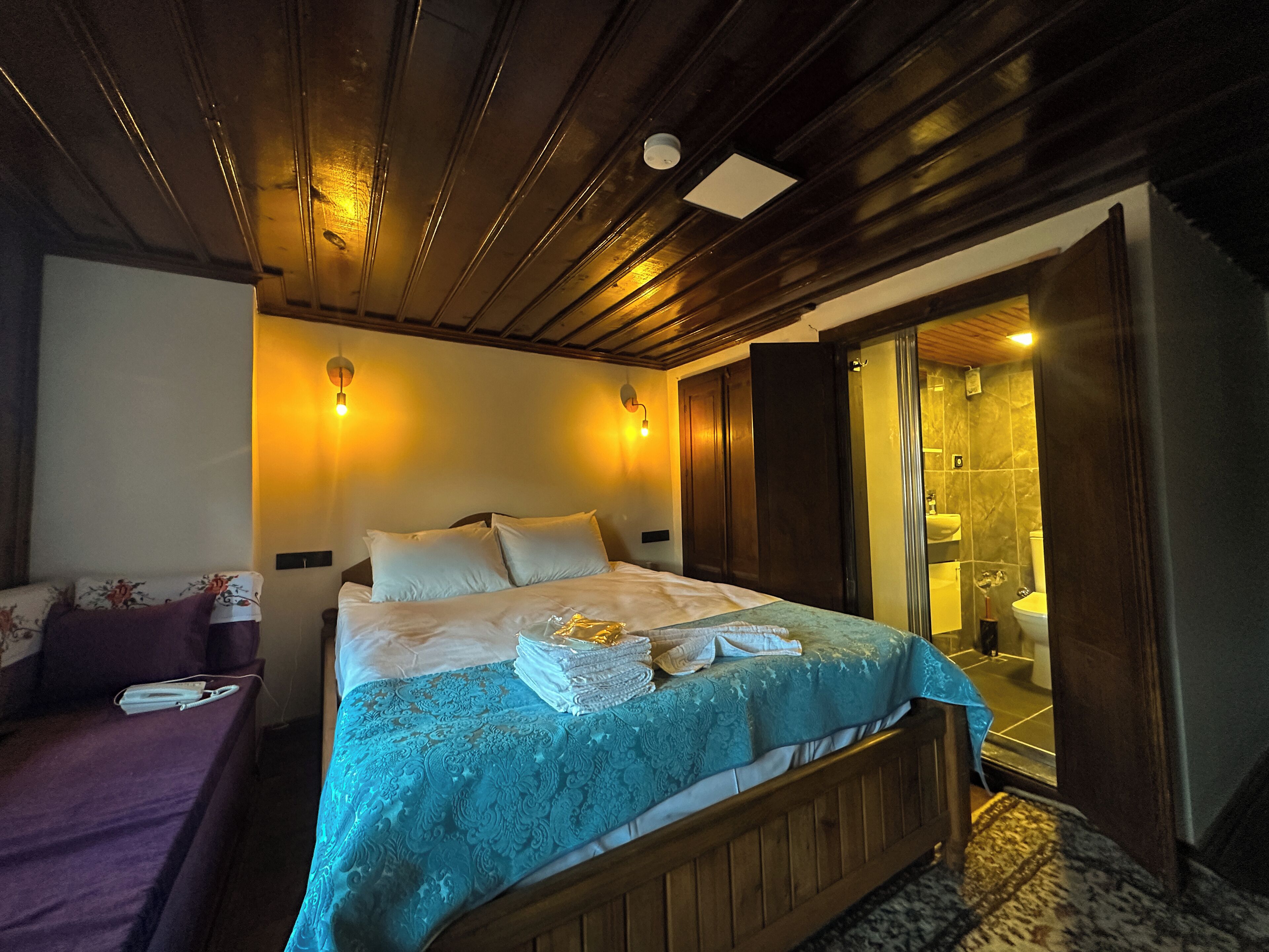 Habitación de hotel con una cama de madera, una mesita de noche, un sofá y un baño con ducha.
